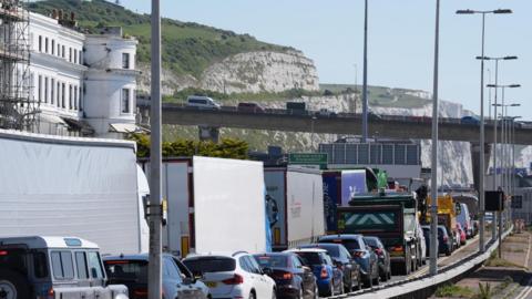 Traffic stationary and queuing along a road near the Port of Dover. Vehicles are also seen on a congested flyover. The white cliffs of Dover can be seen in the background.