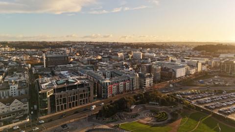 A drone shot showing St Helier from the sky. The sun is low and it gives a golden glow to the buildings packed together. There is a park in the foreground and a car park next to it. 