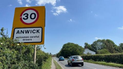 A large orange 30mph sign on the side of the road. Below, it says "Anwick welcomes careful drivers". Two cars, one silver and one blue, can be seen driving on the road away from the camera. There are grass verges and a path on each side of the road. The sky is blue. 