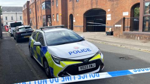 A police car parked up on Bedford Street behind a blue and white cordon. In the background you can see an arched entrance into a brick building where a cordon has been placed.