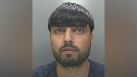 Bilal Dawlatzai in a police custody shot looking at the camera. He has dark hair falling over his eyebrows and facial hair. He is wearing a dark blue T-shirt.