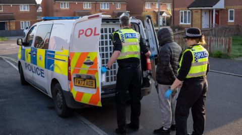 Police putting a man into a police van on a street with houses of light brown brick either side. The man in custody is young and is wearing a grey padded hoodie