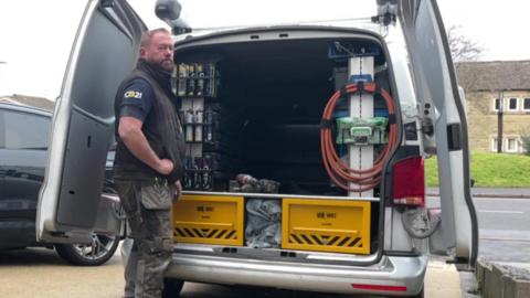 Plumber Andy Cam stands at the back of his silver van, with equipment and piping visible in the back as the two rear doors are open. The vehicle is parked on a driveway, with a car parked next to it. 