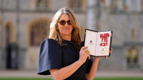Dame Emma Bridgewater has fairish brown shoulder-length hair and is wearing sunglasses and a navy dress. She holds her damehood insignia in a case.