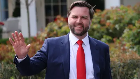 JD Vance wears a blue suit and red tie and is smiling as he waves.