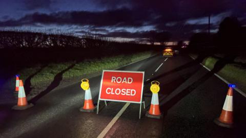 A road closed sign and traffic cones can be seen in the foreground, and emergency service vehicles in the background, of this photograph from the scene.