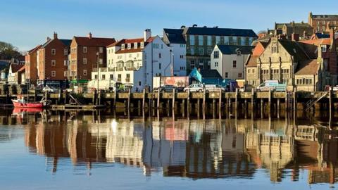 A harbour front of buildings under a blue sky all reflected in the water