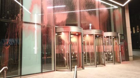 The entrance of a large glass building with three electric doors. Red paint is splashed across all of the windows and doors