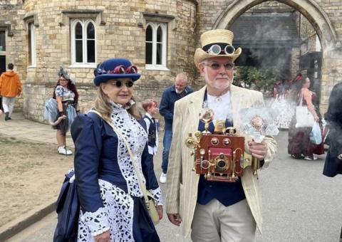 Two people attending the festival, with a man on the right wearing a cream and blue suit with an old Victorian camera and steam coming out of it. He wears a cream top hat and sunglasses. A woman on the left has a blue hat and sunglasses and is wearing a Victorian style blue and white long dress