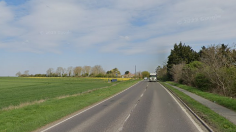 Google image of the A10 during the day. There is grass on either side of the road and a flat field. 