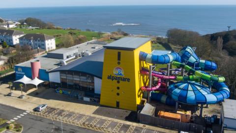 The outside of a water park site, pictured from above. Part of the exterior is bright yellow and reads 'Alpamare'. Water slides can be seen running into the building.
