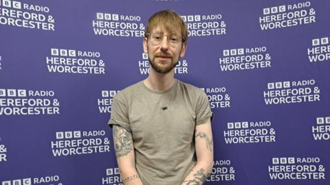 Man wearing round glasses, a grey t-shirt and green necklace is standing in front of a purple wall with the words "鶹ҳ Radio Hereford and Worcester" in the background.