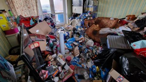 Image shows the inside of Ruth Cookson's living room. It is completely full of empty drinks cans, boxes, carrier bags and other rubbish. There is no floor visible because there is so much covering it. 