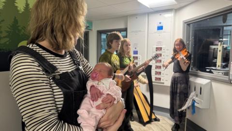 A baby, wearing a pink sleepsuit, sucking a pick dummy, sleeps in her mother's arms. They are listening to the musicians on the hospital ward who are playing a violin, harp and guitars.