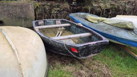 A black, weather-beaten boat can be seen with a red notice stuck to the back. Other boats are pictured either side, sitting atop grass.