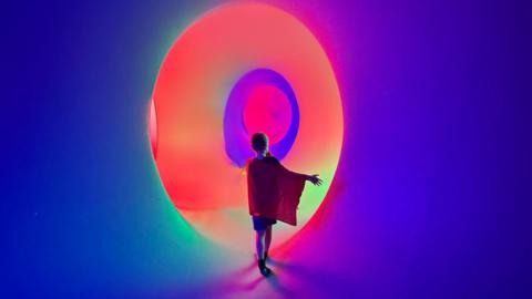 A boy wearing a red cloak and shorts in front of a lit-up installation which is dark blue, green and red and orange. 