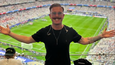 Man wearing black a t-shirt with blue tinted glasses and a handle-bar style moustache with his arms outstretched standing against the backdrop of a crowd-filled American football stadium with his back to the green pitch