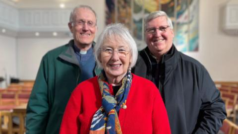 Three people stand inside a church with rows of chairs behind them. In the centre, Sue Gilbert, an elderly woman, smiles at the camera and wears glasses, bright red cardigan and blue scarf. Behind her stand two smiling men, one is wearing a clerical collar.