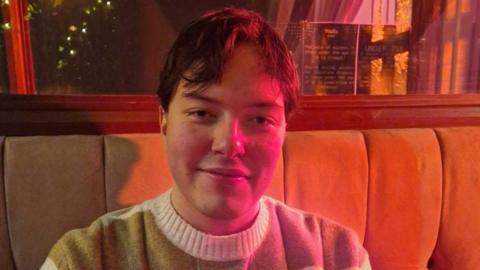 Harrison Harvey is wearing a woolly jumper and has short brown hair. He is sitting on a sofa in a bar/restaurant.