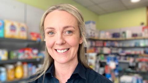 A woman with blonde hair tied back from her face is smiling at the camera. She is wearing a black shirt and you can see shelves of medication etc in the background.