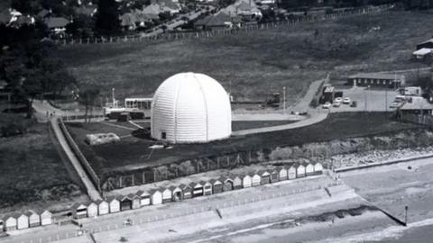 Black and white aerial image of the radar station behind a row of beach huts