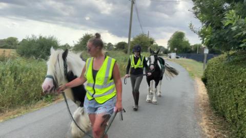 A woman leading a white and black pony. Behind her is a girl leading another black and white pony on a narrow road in between two fields.