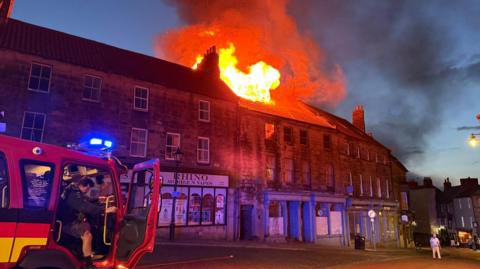 A firefighter opens the door to his fire appliance. Behind, orange flames and smoke can be seen billowing from the roof of a building on a row of terraced shops.
