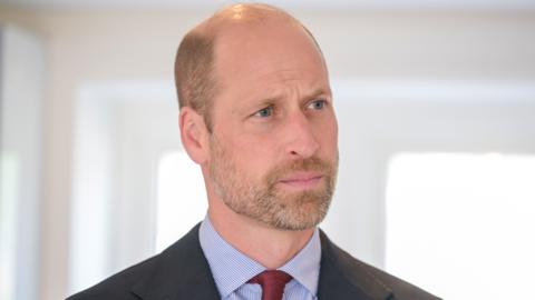 Prince William looking thoughtful during a visit while he wears a black jacket, blue and white striped shirt and dark red tie.