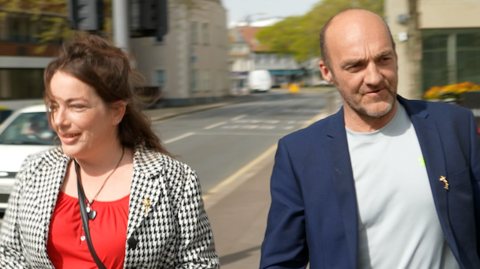 Two people are walking side by side along the street. The person on the left is wearing a black‑and‑white checkered jacket over a red top and carrying a small black bag. The person on the right is wearing a dark blue suit jacket over a light grey shirt. Both individuals have yellow ribbon pins on their clothing. Behind them, a road, parked cars, buildings, and trees are visible.