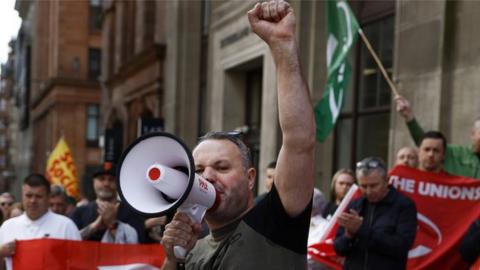 An RMT union member on a strike picket line with his fist in the air holding a megaphone