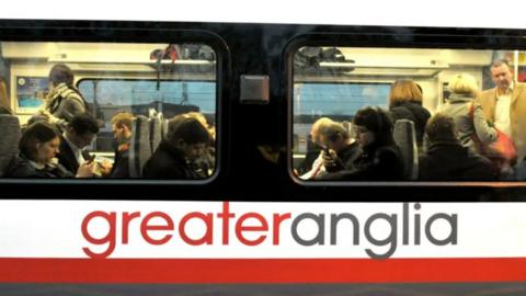 A view of a Greater Anglia train. Passengers can be seen inside the train through large windows. The train operators name has been printed on the side of the carriage.