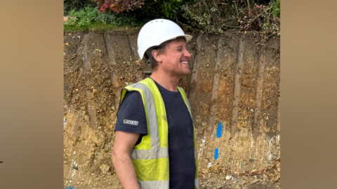 A side shot of Paul, wearing a white builder's hard hat and yellow high vis vest, over a blue Lacoste T-shirt. He is chuckling heartily at something and has his eyes closed.