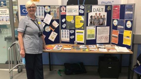 Carolyn has short blonde hair and is standing in front of a table with leaflets and photos on it. She is wearing a blue nurses top and has a black lanyard around her neck