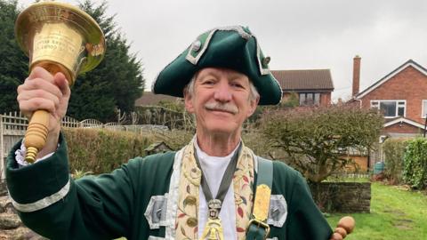 Mr Ward is wearing dark green and silver robes. He has a beige shirt on underneath that has colourful leaves on it. He has a gold medal around his neck, shaped like a bell. He is holding a scroll in one arm, while the other is aloft, holding a large gold bell with a wooden handle. He wears a green and silver hat