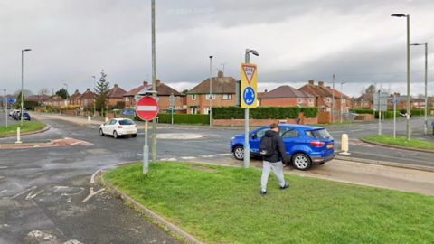 The Holme Lacy Road in Hereford. Cars are crossing a mini-roundabout while someone is walking on a grass verge on the side of the road.