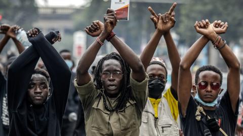Four male protesters, standing in a row, raise their arms in an "X" symbol.