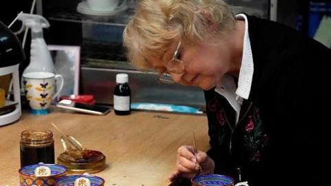 A worker paints the finish on chinaware in a pottery in Stoke-on-Trent. She is leaning forward and using a paintbrush. Paint and china are on the desk in front of her. She is wearing glasses and concentrating.