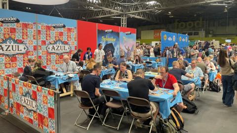A lot of people sitting at tables playing board games at an exhibition. There are people standing around them and advertising boards have been put up next to them.