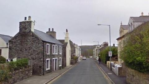 Four Roads in Port St Mary. It has stone built buildings on each side along with green hedges. There are hills in the distance as the road stretches away.
