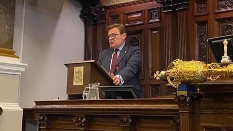 A man in a blue suit with a red tie and glasses, standing behind a wooden lectern in the city council building.