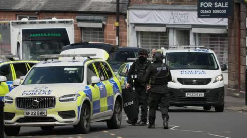 Two armed police officers in bullet-proof vests and wearing black helmets stand next to three police cars and a police van on the street
