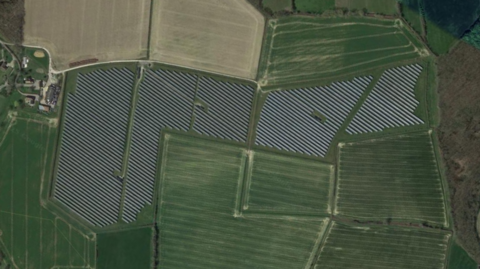 A bird's eye view of a solar farm. The farm includes lots of black solar panels surrounded by green and cream-coloured fields