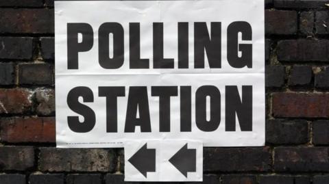 A white paper sign with the words 'polling station' in black letters, with two black arrows pointing left. The sign is stuck to a dark brick wall