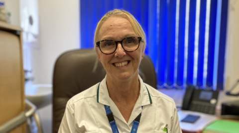 A woman with blonde hair and glasses looking at the camera and smiling. She is wearing a white uniform and a blue lanyard. The background is blurred but she is sat in a brown chair in an office