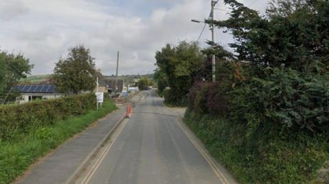 The road in Nanstallon with trees on either side. There is a pavement and a road down the middle.