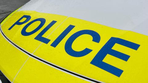 The front of a police vehicle with the word 'Police' on the bonnet in blue. The car is yellow and white.