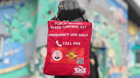 A person stood in front of a graffitied wall holding a red square kit with a handle. It reads 'Public access bleed control kit. Emergency use only. Call 999'. It has a QR code and several logos. 