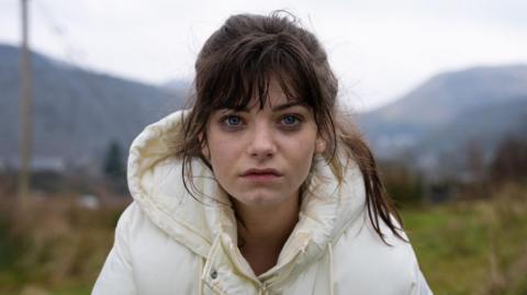A young woman with brown hair in a ponytail and wearing a white puffer coat stares at the camera with a sad expression