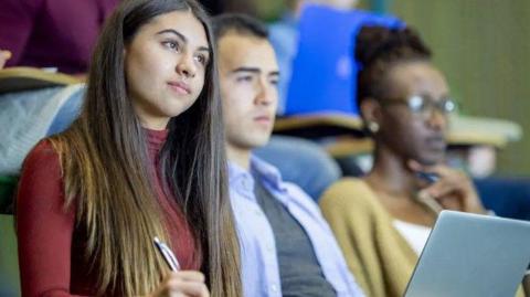 Three students at a lecture. One is dressed in a red shirt holding a pen, while the other has a laptop open. 