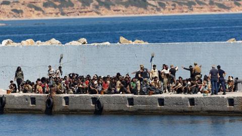 A large crowd of migrants waits on a concrete jetty
